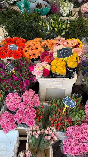 A colorful floral display at a flower stall featuring a variety of fresh flowers arranged in different containers. In the foreground, there are clusters of pink roses in a white basket, alongside vibr